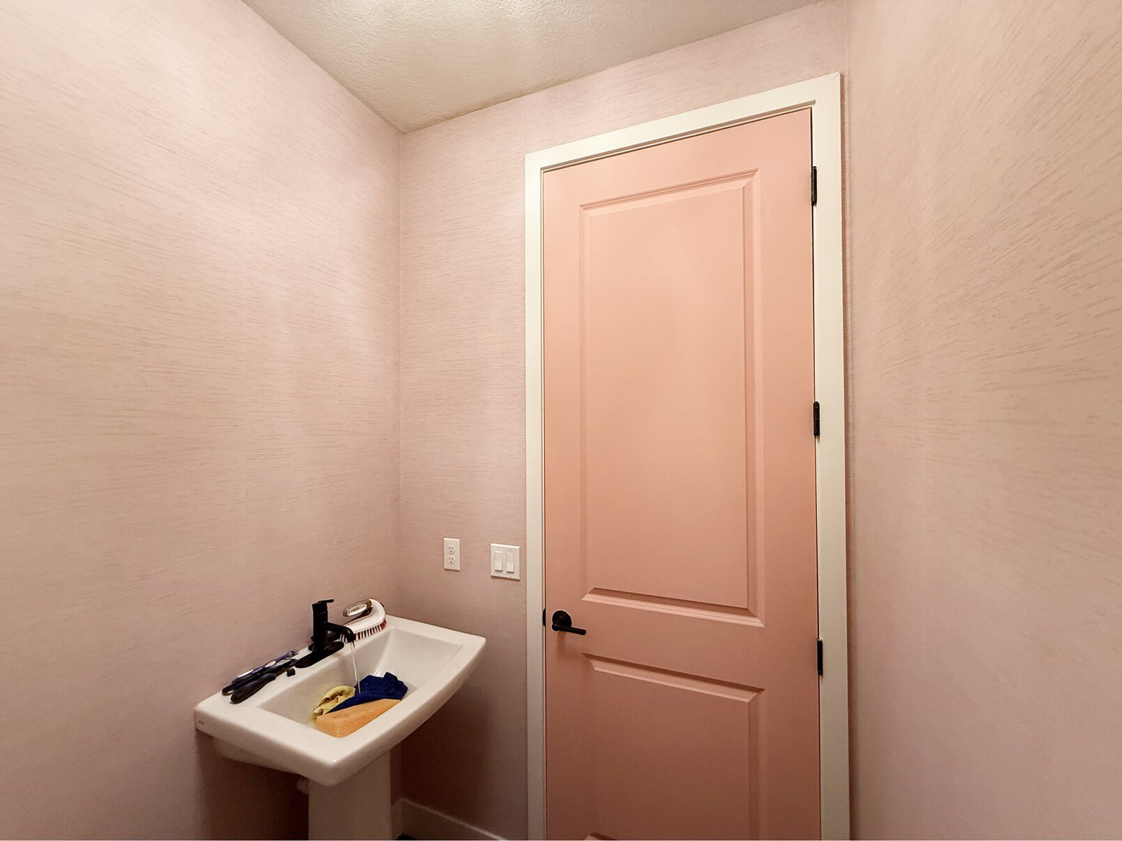 Powder room with textured blush pink wallpaper, white pedestal sink, and peachy-pink paneled door at Green Valley Ranch Golf Club in Colorado